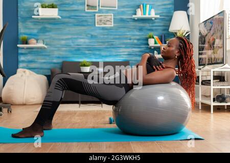 Fit sporty strong black woman working out core abdominals doing crunch exercise using swiss stability ball, on yoga mat in home living room dressed in sport wear and concentration on corect breathing. Stock Photo