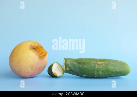 Rotten, bruised, stale or bad apple and cucumber isolated in blue background. Food waste and poor agricultural crop quality concept. Stock Photo