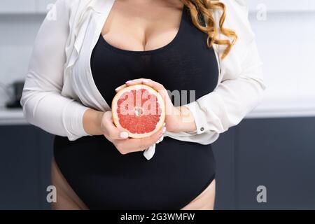 Close up curvy body young woman holding a cut half of a fresh grapefruit in hands standing on the modern kitchen at home with lo Stock Photo