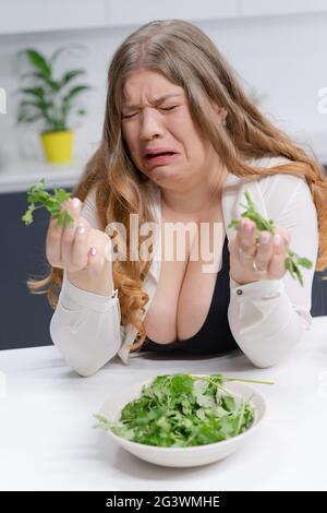 Fat girl not happy holding fresh salad casting. Curvy body young woman with long blond hair sitting on modern kitchen. Dieting a Stock Photo