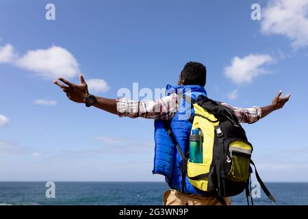 Fit afrcan american man wearing backpack hiking spreading arms on the coast Stock Photo