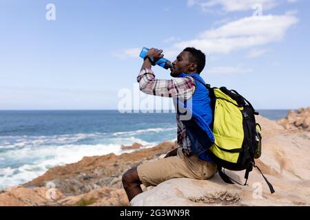 Fit afrcan american man wearing backpack resting drinking water on the coast Stock Photo