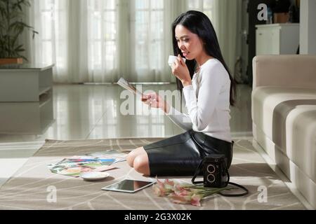 Content young Vietnamese photographer with black long hair sitting on floor in modern living room and viewing photos while drinking coffee Stock Photo