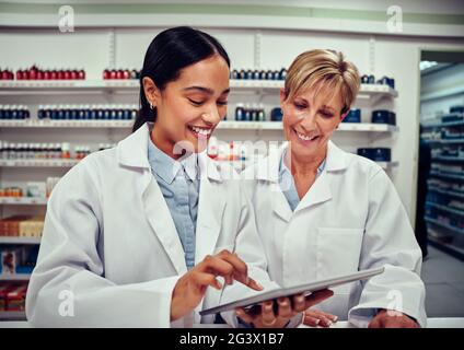 Young woman using a digital tablet and showing something to her senior colleague wearing labcoat while working in pharmacy Stock Photo