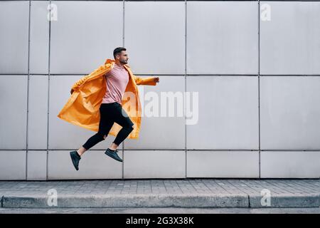Trendy young man in yellow raincoat jumping in the air on wall street urban background Stock Photo
