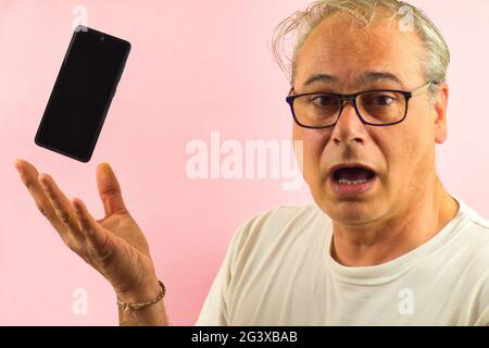 mature man with glasses and grey hair shows surprised a mobil with outstretched hand on pink background Stock Photo