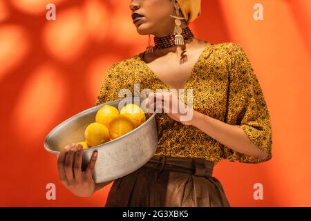 partial view of african american woman in yellow blouse holding metal bowl with lemons on orange Stock Photo