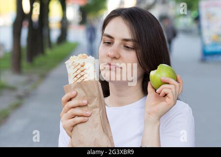 Young beautiful brunette woman on a diet chooses food - what to eat an apple or fast food. Stock Photo