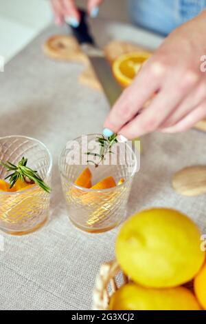 Female hand put rosemary in glasses, Woman preparing, making citrus and rosemary fresh lemonade in glass on a white table at home, summer drink, detox Stock Photo