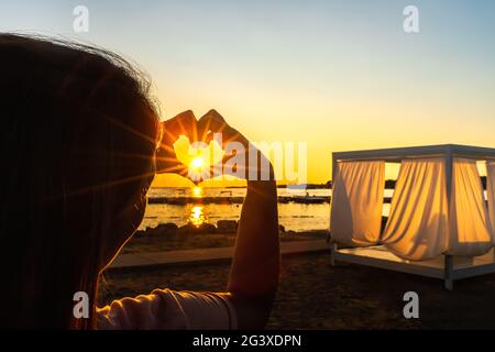 Woman showing with her hands heart witch trough shines Sunstar with sunrays, at beautiful sunset on Stock Photo