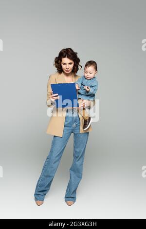 full length of businesswoman in blazer holding clipboard and baby boy in arms on grey Stock Photo