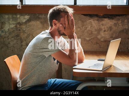 Side view of upset male student with hands on face tormented by work. Young man in grey T-shirt is working on laptop in cafe. Concept of remote studying. Stock Photo