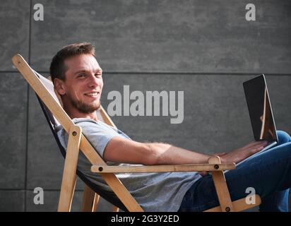 Smiling male sitting on wooden chair and typing on laptop. Young man working on laptop outdoor under sunny weather. Concept of comfortable workplace. Stock Photo