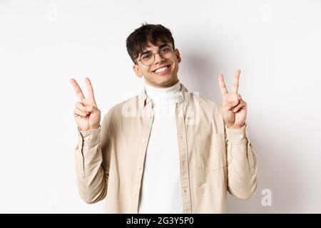 Handsome hipster guy in glasses, smiling with white teeth, showing peace or victory gesture, standing upbeat on white background Stock Photo