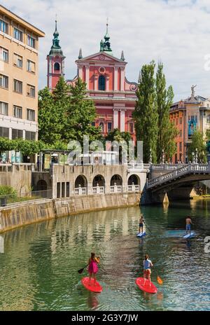 Ljubljana, Slovenia.  Group of young people paddling down the Ljubljanica River on paddle surf boards.  Baroque Franciscan Church of the Annunciation Stock Photo