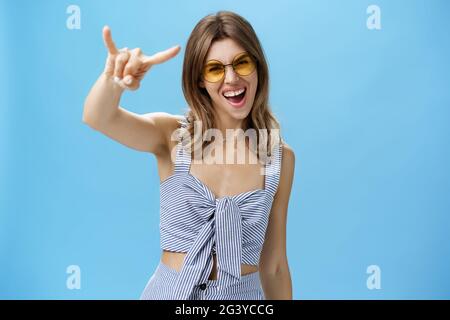 Waist-up shot of confident and daring charming young woman in matching trendy clothes yelling yeah showing rock-n-roll gesture a Stock Photo