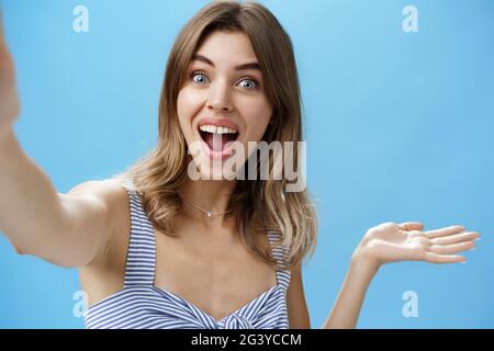 Waist-up shot of charismatic upbeat enthusiastic cute female blogger raising hand over copy space and holding camera as if recor Stock Photo