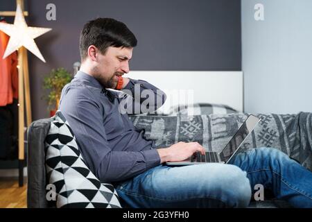 Tired strained man suffering from neck pain while working at computer. Male touchingly massages neck, puts on neck collar, sitti Stock Photo