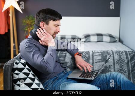 Tired strained man suffering from neck pain while working at computer. Male touchingly massages neck, puts on neck collar, sitti Stock Photo