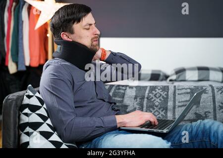 Tired strained man suffering from neck pain while working at computer. Male touchingly massages neck, puts on neck collar, sitti Stock Photo