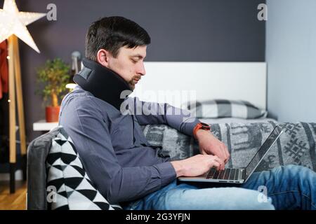 Man employee after accident working at home behind laptop and feels pain in neck, uses black neck collar support. Male with neck Stock Photo