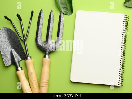 Open notebook with blank white sheets and various gardening tools with wooden handles on green background, flat lay Stock Photo