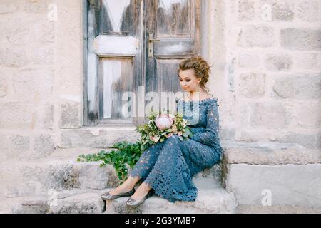 A bride in a stylish blue dress with a bouquet in her hands sits on the steps near an old wooden door Stock Photo