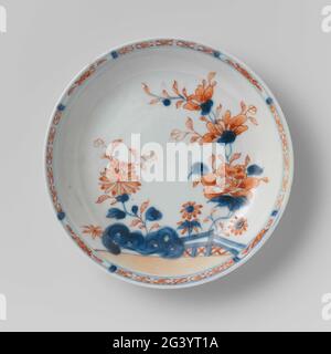 Saucer With Flower Sprays Behind a Rock and Fence. Dish of porcelain, painted in underglaze blue and on the glaze red and gold. On the flat of the dish large, blooming plants (peony, chrysanthemum, aster) behind a rock and a fence; The edge with napkin interspersed with a flower branch in a cartouche. Chinese Imari. Stock Photo