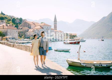 A man and a woman in sunglasses walk hugging each other on the pier near the boats in old town of Perast Stock Photo