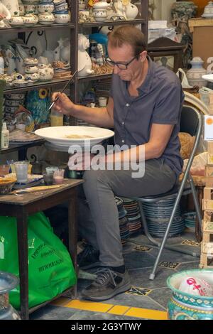 An artisan workshop in Vietri - Italy. A craftsman is painting a clay plate. Handmade souvenirs. Many pottery objects at the display shelfs. Stock Photo