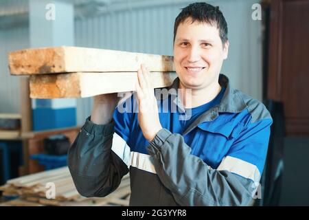 A young man stands in a carpentry shop and holds wooden bars on his shoulder. Stock Photo