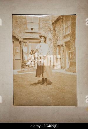 Authentic vintage sepia photograph of smiling young woman carrying flowers standing in front of house. Concept of nostalgia, lifestyle, historical Stock Photo