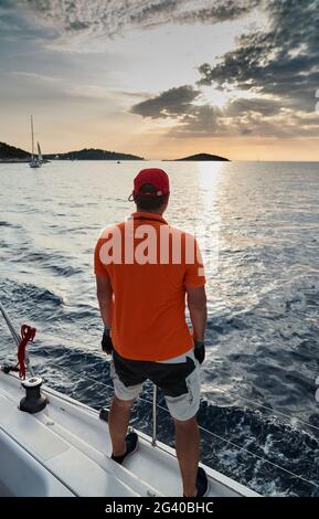 The participant of a sailing regatta is on the edge of the boat, he enjoys a victory and a sunset, he is dressing in t-shirt of Stock Photo