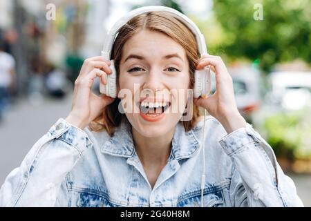 Portrait of a cheerful stylish young woman wearing denim jacket standing isolated over city background, listening to music with headphones Stock Photo
