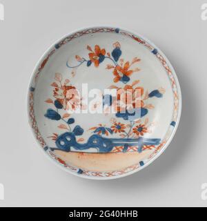 Saucer With Flower Sprays Behind a Rock and Fence. Dish of porcelain, painted in underglaze blue and on the glaze red and gold. On the flat large, blooming plants (peony, chrysanthemum, aster) behind a rock and a fence; The edge with napkin interspersed with a flower branch in a cartouche. Chinese Imari. Stock Photo