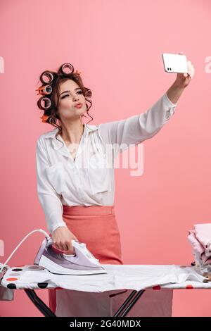 Against a pink background, a beautiful housewife with a hair curler works clothes on the ironing board and takes a selfie on the phone. Cute girl doin Stock Photo