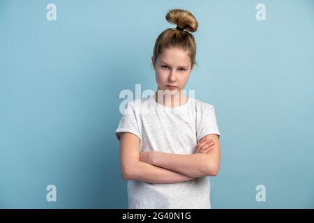 Young teen girl folded her arms across her chest and looks to camera on a blue background. The girl angrily looks isolated on the copy space. Stock Photo