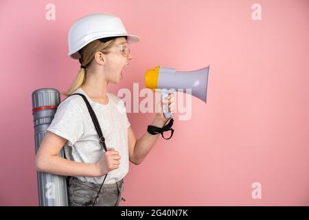 Portrait of a teenage girl in a helmet, a loudspeaker in his hands and a top hat on his back. Girl isolated on a pink background, shouting through a m Stock Photo