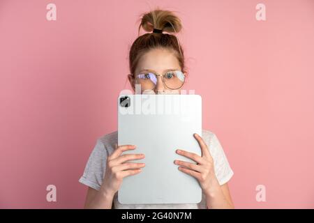 Attractive, blond girl with a tablet and glasses on a pink background. Cute teenager uses a tablet, she covered her face with a tablet, only her eyes Stock Photo