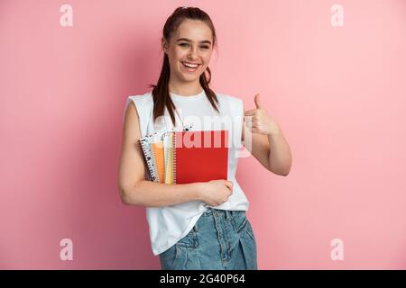 Cute, positive girl with books in her hands shows thumbs up, okay sign. Female student with note and books on isolated pink background., Cute, positiv Stock Photo