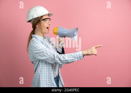 Young pretty woman over isolated pink background shouting through megaphone. Beautiful girl in a helmet and construction glasses shows how to do the j Stock Photo