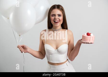 Cheerful, positive lady licks, wants to eat an appetizing cake. In her hand a girl holding balloons, posing on a white background, Cheerful, positive Stock Photo