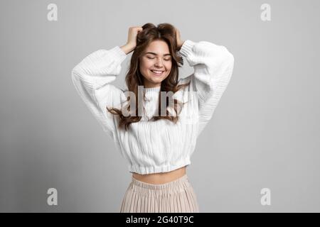 Charming young lady touches her dark hair on a gray background. Cute girl closed her eyes, posing beautifully against the background of an empty wall. Stock Photo
