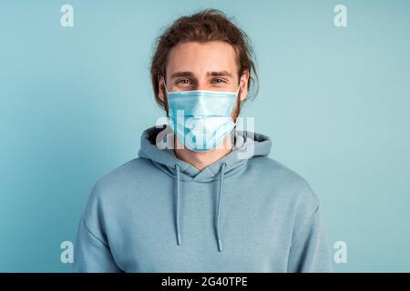 Portrait of young handsome man with medical mask standing, self defense concept. Indoor studio shot isolated on blue background. Stock Photo