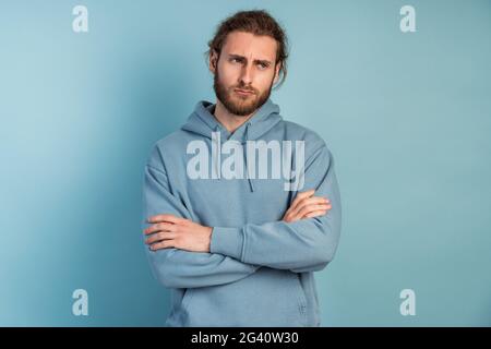 Photo of an upset man in a hooded sweater, he crossed his arms over his chest. Man posing on a blue background. Stock Photo