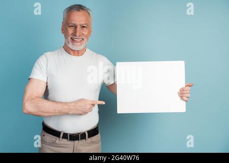 Smiling, pleasant, senior man holds a white piece of paper, points to it. Isolated on blue background man holding blank white piece of paper, copy spa Stock Photo