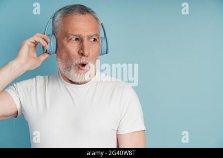 Surprised man in headphones posing on blue wall background, isolated. Positive senior man listening to music on headphones, Surprised man in headphone Stock Photo