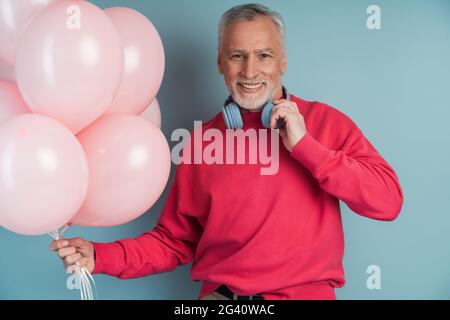 A smiling, older bearded man wears headphones, holding balloons in his hand. Senior man posing on blue background, A smiling, older bearded man wears Stock Photo