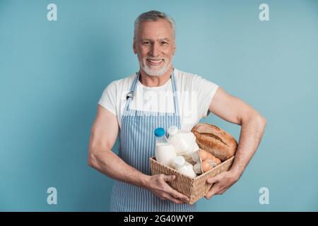 Smiling man with gray hair and beard holding a food basket. Cute grandfather posing on a blue background. Stock Photo