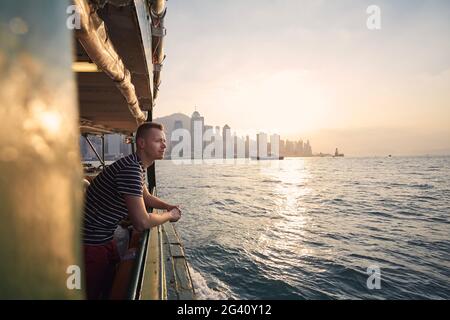 Pensive man looking from ferry boat against urban skyline st beautiful sunset. Tourist in Hong Kong. Stock Photo